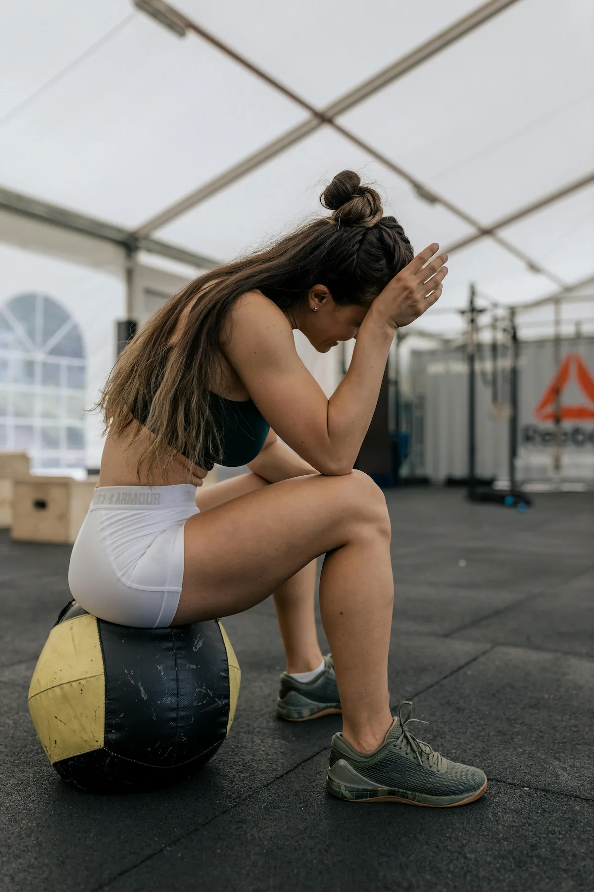 A woman in athletic wear sits on a medicine ball with her head bowed and hand on her forehead, appearing exhausted after a workout in a gym setting.