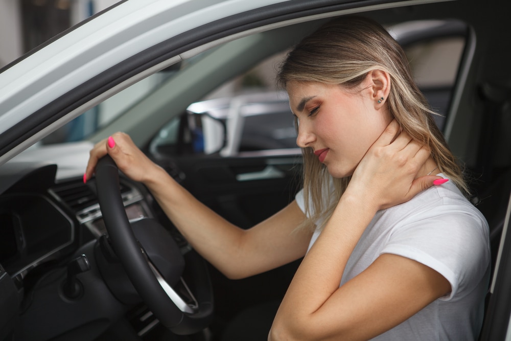 A young woman in a white t-shirt sits in a car, holding the steering wheel with her left hand and her right hand on the back of her neck, indicating neck pain or discomfort.