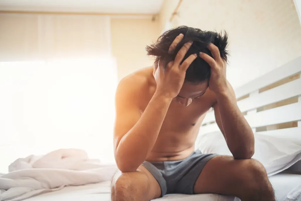 A shirtless man with disheveled hair sits on a bed, hunched over with his hands gripping his head, appearing to have a headache.