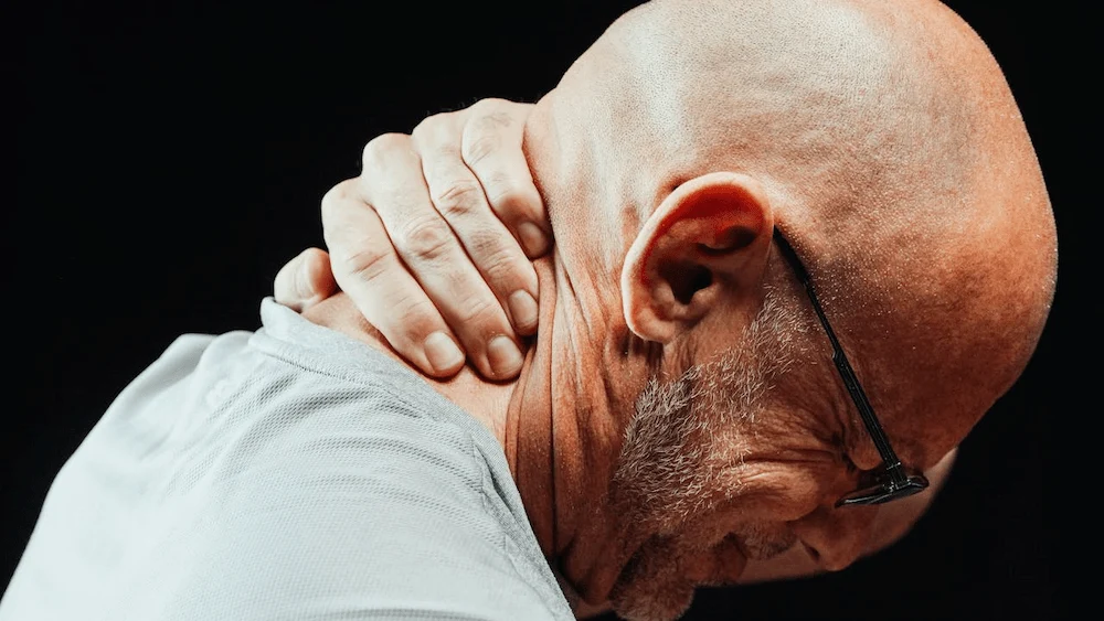Side profile of a bald person with a beard and glasses, holding their neck with their hand, indicating pain or discomfort.